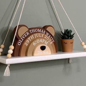 May include: A wooden bear-shaped rainbow shelf with the text "Oliver Thomas Waite 29th July 2024 6:04PM 7lb 4oz" hanging from a white shelf with tassels. A small potted succulent plant is on the shelf.