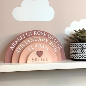 May include: A wooden rainbow-shaped sign in pink and peach tones, inscribed with "ARABELLA ROSE DRAPER", birth details, and weight. A heart shape is in the center. The sign is on a white shelf with a decorative plant.