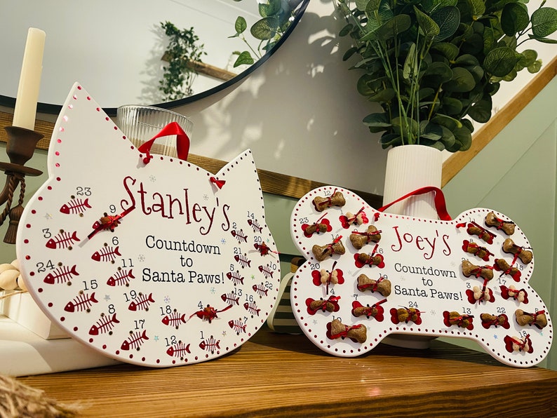 May include: Two white wooden Christmas countdown calendars, one shaped like a cat and the other like a bone. The cat calendar has red fish bones with numbers on them and says "Stanley's Countdown to Santa Paws!" The bone calendar has red bones with numbers on them and says "Joey's Countdown to Santa Paws!"