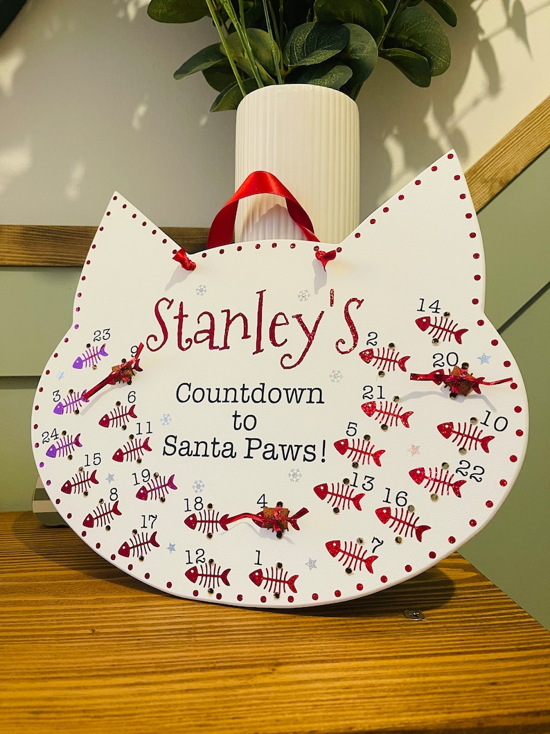 May include: White wooden cat-shaped countdown calendar with red glitter fish bones and the text "Stanley's Countdown to Santa Paws!"
