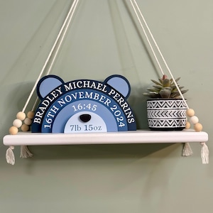 May include: A blue and white wooden rainbow baby birth announcement with the text "Bradley Michael Perrins, 16th November 2024, 16:45, 7lb 15oz". The rainbow is hanging from a white string and is displayed on a white shelf with tassels.