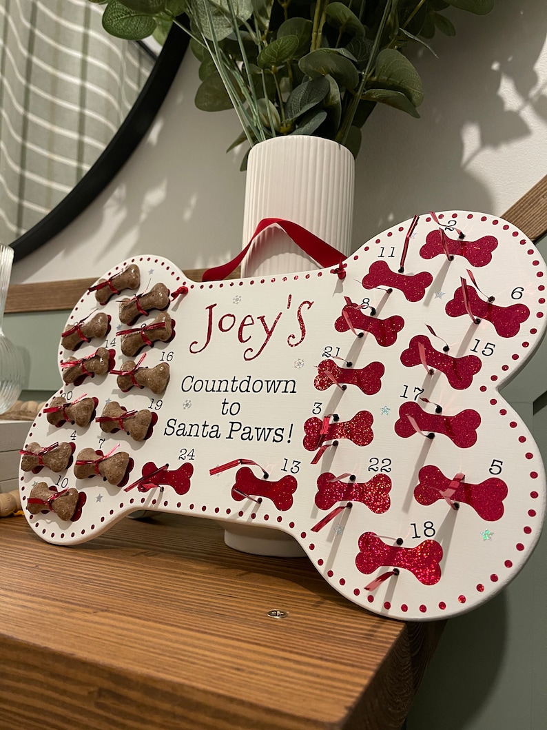 May include: White wooden bone-shaped advent calendar with red glitter and red ribbon. The calendar is decorated with paw prints and stars. The text "Joey's Countdown to Santa Paws!" is written on the calendar.