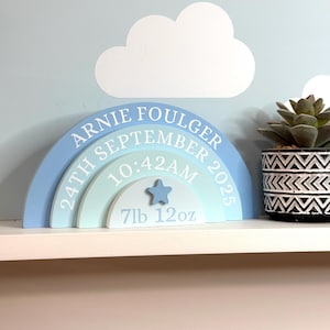 May include: A wooden rainbow-shaped nursery sign in shades of blue and teal. The text reads ARNIE FOULGER, 24TH SEPTEMBER 2025, 10:42AM, and 7lb 12oz. A small blue star is attached to the centre. The sign is displayed on a white shelf.