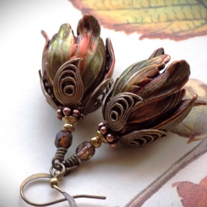 Pode incluir: Um par de brincos com um design de flores em bronze e verde. As flores s&atilde;o detalhadas com filigrana intrincada e t&ecirc;m um visual vintage. Os brincos s&atilde;o suspensos por um fio de bronze com uma conta de vidro marrom escuro.