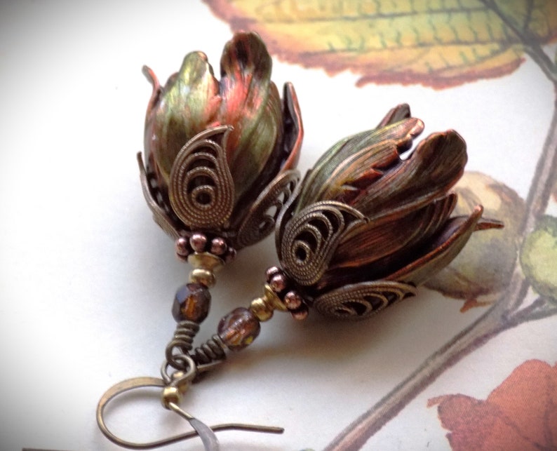 Pode incluir: Um par de brincos com um design de flores em bronze e verde. As flores s&atilde;o detalhadas com filigrana intrincada e t&ecirc;m um visual vintage. Os brincos s&atilde;o suspensos por um fio de bronze com uma conta de vidro marrom escuro.