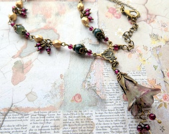 Op de afbeelding: Een handgemaakte ketting met een bloemenhanger en diverse kralen. De ketting heeft een bronzen ketting met een bloemvormige hanger in roze en witte tinten. Het bevat ook groene en gouden kralen en kleine rode kralen.