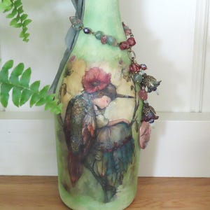 May include: A light green decorative glass bottle featuring a printed illustration of a figure with a red flower headpiece and bird-like wings. The bottle is adorned with a grey ribbon tied at the neck and a beaded chain with floral metal charms.