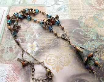 Pode incluir: Um colar delicado com um pingente floral. O colar apresenta uma mistura de contas de vidro azul, verde e laranja, e uma corrente de bronze. O pingente é uma flor estilizada com um esmalte azul e verde.