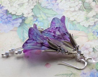 Pode incluir: Brincos florais roxos e azuis com detalhes prateados e contas de pérola. Os brincos têm a forma de flores com um efeito marmorizado.
