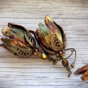 Pode incluir: Um par de brincos pendentes com um design de flores em cobre e verde. As flores s&atilde;o detalhadas com filigrana e t&ecirc;m um visual vintage. Os brincos s&atilde;o acentuados com contas marrons e iridescentes.