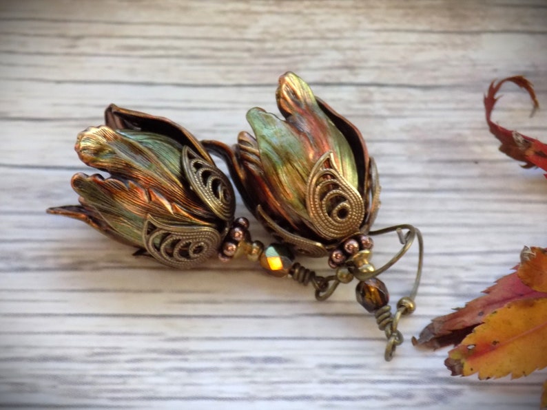 Pode incluir: Um par de brincos pendentes com um design de flores em cobre e verde. As flores s&atilde;o detalhadas com filigrana e t&ecirc;m um visual vintage. Os brincos s&atilde;o acentuados com contas marrons e iridescentes.