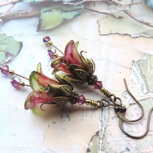 May include: A pair of handcrafted earrings featuring delicate, flower-shaped pendants. The earrings showcase pink and green floral designs with gold-colored accents and purple crystal beads. The ear wires are a dark bronze color.