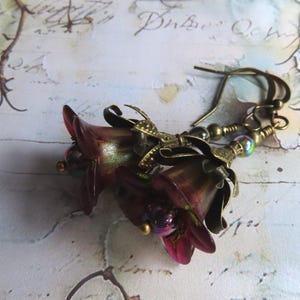 May include: A pair of drop earrings featuring dark burgundy, bell-shaped flower charms with iridescent centers. The flowers are accented with antique brass-toned metal caps and small, multi-colored metallic beads, suspended from matching brass ear wires.