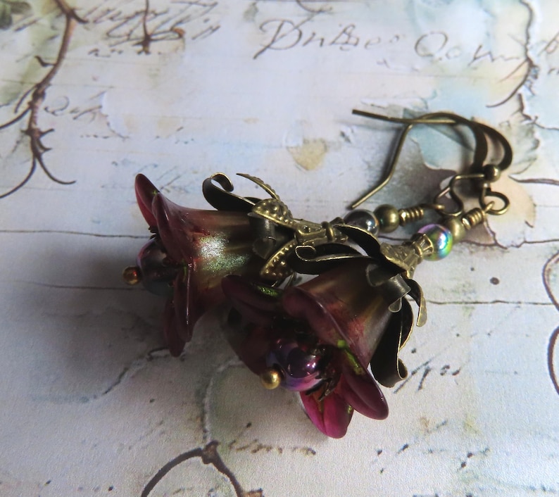 May include: A pair of drop earrings featuring dark burgundy, bell-shaped flower charms with iridescent centers. The flowers are accented with antique brass-toned metal caps and small, multi-colored metallic beads, suspended from matching brass ear wires.