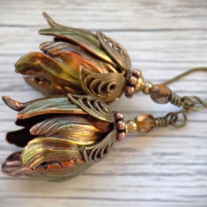 Pode incluir: Um par de brincos com um pingente em forma de flor feito de cobre e metal dourado iridescente. Os brincos s&atilde;o adornados com pequenas contas marrons e transparentes e t&ecirc;m um fecho de gancho.