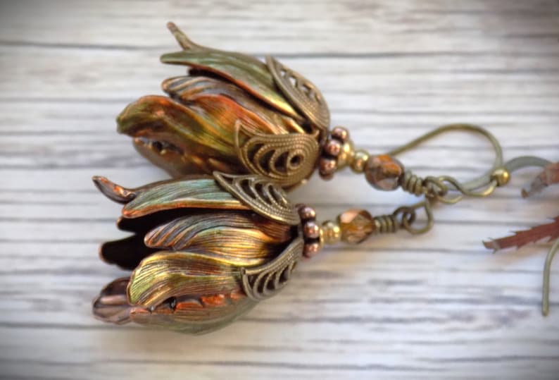 Pode incluir: Um par de brincos com um pingente em forma de flor feito de cobre e metal dourado iridescente. Os brincos s&atilde;o adornados com pequenas contas marrons e transparentes e t&ecirc;m um fecho de gancho.