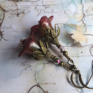 May include: A pair of dangle earrings featuring bell-shaped flower beads in a gradient of deep red and gold. Each flower is accented with an antique brass-toned metal cap, small iridescent glass beads, and a curved brass ear wire hook.
