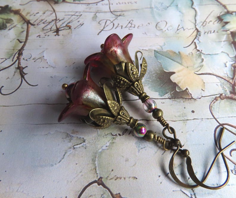 May include: A pair of dangle earrings featuring bell-shaped flower beads in a gradient of deep red and gold. Each flower is accented with an antique brass-toned metal cap, small iridescent glass beads, and a curved brass ear wire hook.