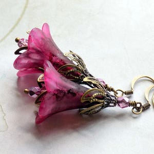 May include: A pair of dangle earrings featuring two pink glass flower charms with bronze metal accents and pink crystal beads.
