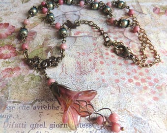 May include: A vintage-style necklace featuring a pink and bronze color scheme. The necklace has a floral pendant and is adorned with pink beads and bronze-colored chain links. The background is a floral pattern.