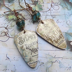 Könnte beinhalten: Ein Paar Ohrhänger mit schildförmigen Anhängern und Etiketten im Vintage-Stil. Ein Etikett trägt die Aufschrift Sylph's Ethereal Mist, das andere Luminous Pixie Dust. Jeder Ohrring hat eine facettierte türkisfarbene Perle und eine bronzene Kappe.