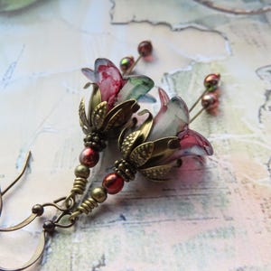 May include: A pair of floral earrings with a vintage aesthetic. The earrings feature translucent, pink and green flower-shaped pendants with brass-coloured metal accents and red beads. The ear hooks are a bronze colour.
