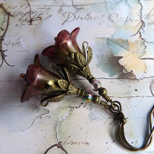 May include: A pair of drop earrings featuring two bell-shaped, deep magenta glass flower beads encased in antique brass-toned metal caps. The earrings include small iridescent glass beads and are finished with brass-toned metal ear wires.