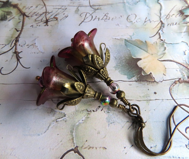 May include: A pair of drop earrings featuring two bell-shaped, deep magenta glass flower beads encased in antique brass-toned metal caps. The earrings include small iridescent glass beads and are finished with brass-toned metal ear wires.