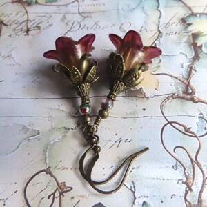 May include: A pair of dangle earrings featuring translucent magenta and amber bell-shaped flower beads. Each flower is set in an antique brass-toned metal calyx, accented with a small iridescent bead on a brass wire, finished with a brass hook ear wire.