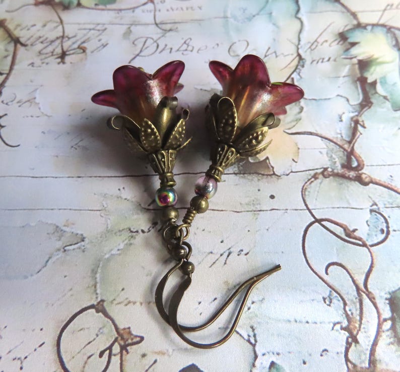 May include: A pair of dangle earrings featuring translucent magenta and amber bell-shaped flower beads. Each flower is set in an antique brass-toned metal calyx, accented with a small iridescent bead on a brass wire, finished with a brass hook ear wire.