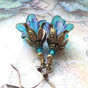 May include: A pair of handcrafted earrings featuring blue and purple flower-shaped pendants. The pendants are accented with bronze-colored metalwork and turquoise beads. The earrings have a vintage aesthetic and are suspended from bronze-colored ear wires.
