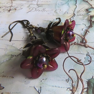 May include: A pair of dangle earrings featuring dark magenta flower-shaped beads with metallic green edges. Each flower has a central iridescent purple bead and is attached to a bronze-toned metal hook with small matching accent beads.