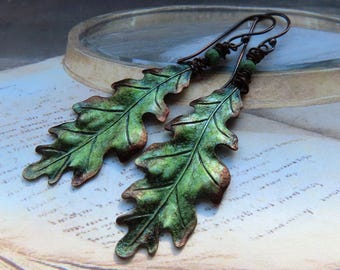Pode incluir: Um par de brincos artesanais com desenhos detalhados de folhas de carvalho. As folhas são pintadas em tons de verde com detalhes em castanho, suspensas em ganchos de metal escuro. Os brincos têm uma estética rústica inspirada na natureza.