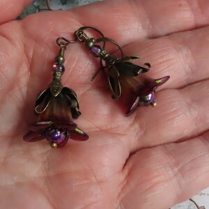 May include: A pair of dangle earrings featuring dark purple and brown bell-shaped flower beads with antique brass-toned metal caps and small iridescent purple beads at the center, suspended from matching antique brass-toned metal earring hooks.