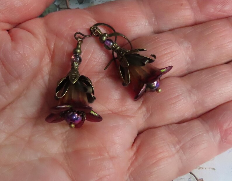 May include: A pair of dangle earrings featuring dark purple and brown bell-shaped flower beads with antique brass-toned metal caps and small iridescent purple beads at the center, suspended from matching antique brass-toned metal earring hooks.