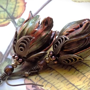 Pode incluir: Um par de brincos com um design de flor de cobre e verde com detalhes intrincados de filigrana. Os brincos s&atilde;o suspensos por uma corrente longa e fina com uma conta marrom.
