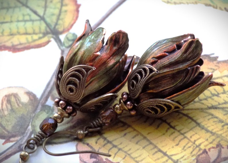 Pode incluir: Um par de brincos com um design de flor de cobre e verde com detalhes intrincados de filigrana. Os brincos s&atilde;o suspensos por uma corrente longa e fina com uma conta marrom.