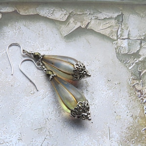 May include: A pair of drop earrings featuring elongated, translucent, pale yellow teardrop-shaped beads with vertical ridges. Each bead is capped with an intricate, silver-toned metal filigree at the base and accented with a small, dark metallic bead near the silver-toned ear wire hook.