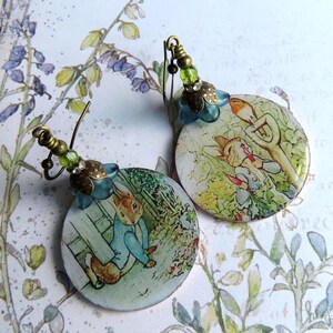May include: Dangle earrings with Peter Rabbit illustrations. Each circular charm depicts a scene from the story, complemented by blue flower beads and green accents. The earrings are a charming accessory.