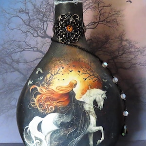 May include: A decorative glass bottle painted in black with a central illustration of a person with long flowing orange hair riding a white horse. The bottle neck features a black filigree metal ornament with an amber stone and a black chain.