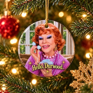 Op de afbeelding: Een ronde keramische kerstornament met een vrouw met rood haar en blauwe oogschaduw die een lichtblauwe telefoonhoorn vasthoudt. Ze draagt een paarse top en een kralenketting. De tekst Hello, Durwood... staat in het geel onderaan afgebeeld.