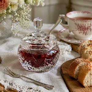 May include: A clear, cut-glass jam jar with a lid and a silver spoon, filled with red jam. A teacup with a floral design and bread slices are nearby, suggesting a breakfast or tea setting.