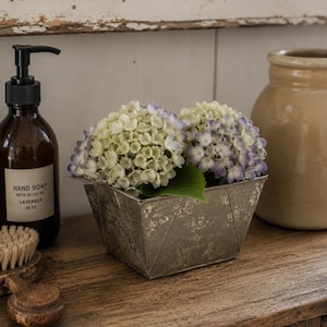 May include: Two hydrangea flower heads with pale green and light purple petals sit in a square, distressed metallic planter. To the left is a dark amber glass bottle with a black pump dispenser and a label reading HAND SOAP WITH OLIVE OIL LAVENDER 16 OZ.