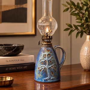 May include: A blue ceramic oil lamp with a raised tree relief design, featuring a brass burner mechanism and a clear glass chimney. The lamp has a curved handle on the side and a lit wick, creating a warm glow within the transparent glass globe.