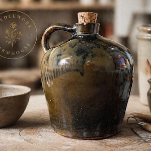 May include: A glazed ceramic jug with a handle and a cork stopper. The jug has a dark brown and green mottled finish. The image also includes a small bowl and other pottery items, suggesting a rustic, farmhouse theme. The jug is approximately 10 inches tall.