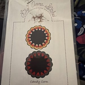 May include: A printed craft pattern sheet titled Lily Anna Stitches featuring a lily illustration at the top. It displays two circular designs: one with a candy corn border and one with a watermelon slice border. The text reads Candy Corn & Summer Slices Pattern LAS200.