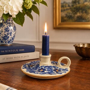May include: A round, cream-colored ceramic candle holder with a blue floral pattern and a side handle. A single dark blue taper candle is inserted into the center and is lit, featuring a bright flame. The holder has a smooth, glazed finish.