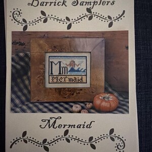 May include: A printed pattern booklet titled Barrick Samplers Mermaid featuring a cross-stitch design of a mermaid with the letters Mm and the word Mermaid. The cover displays a framed version of the needlework next to small pumpkins.