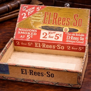 May include: An open El-Rees-So cigar box with red and gold lettering. The box reads "2 for 5¢" and "Millions Smoked at 5¢". The box is made of cardboard and has a vintage aesthetic.