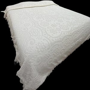 May include: A cream-colored bedspread featuring a textured, raised floral and scrollwork pattern across the entire surface. The edges of the rectangular textile are finished with a decorative fringe detail along the sides and bottom hem.